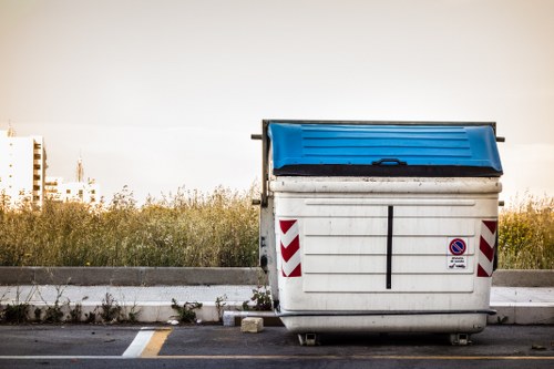 A large commercial skip bin with a blue lid positioned on an asphalt surface in an outdoor area. The bin is predominantly white with red and white striped warning reflectors on its sides and a small no-parking sign affixed to the front. The background features a grassy verge with some overgrown plants and a distant view of a city skyline, including tall buildings. The scene is lit by natural daylight, suggesting a clear or slightly overcast day. The surrounding environment indicates a typical outdoor setting for rubbish collection or waste disposal, consistent with services offered by Commercial Waste Barnet in the Barnet area, accessible via postal code in North London.