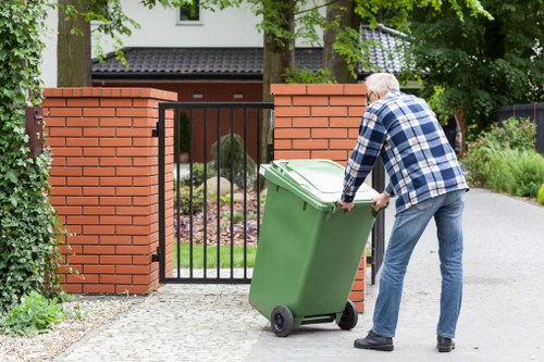 A senior man wearing a blue and white checked shirt and blue jeans is seen emptying a large green wheeled rubbish bin into an open entrance gate of a residential property. The gate, made of black metal bars, is flanked by red brick pillars, and the area around the entrance features a gravel pathway. Behind the gate, there is a well-maintained garden with lush greenery and flowering plants, along with a modern house partially visible in the background. The scene takes place outdoors during daylight, with natural light illuminating the area. The image demonstrates rubbish removal activity typical for domestic waste collection services, aligning with the context of commercial waste removal in Barnet serviced by Commercial Waste Barnet.