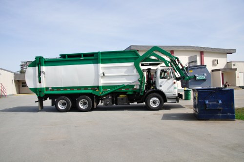 A large commercial waste collection truck from Commercial Waste Barnet is parked on a paved area in front of a modern residential building. The truck has a white body with bright green detailing along the top and sides, and features a hydraulic lifting arm at the rear, used for loading and unloading waste containers. Visible on the front side of the truck is a cab with side mirrors, and the truck's wheels are black with silver hubcaps. To the right of the truck, there are two large waste bins: a dark blue bin positioned on the pavement near the truck, and another bin of similar size placed in the driveway of the building, which has a grey roof and white exterior walls. The background includes a clear sky and glimpses of other buildings, indicating a suburban or urban environment within Barnet. The overall scene demonstrates professional rubbish collection activity relevant to local commercial waste removal services in the Harrow post code area.