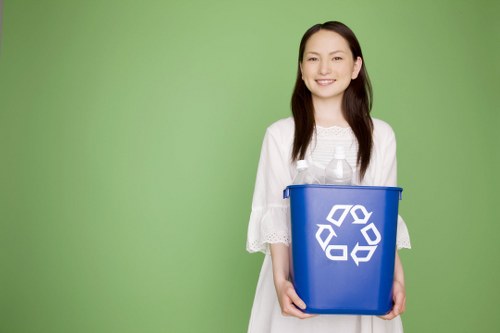 A woman with long dark hair, dressed in a white top with lace detailing on the sleeves, is standing outdoors against a bright green background, smiling at the camera. She is holding a blue plastic recycling bin with a white recycling symbol printed on the front, which appears to be empty and in good condition. The scene is well-lit, with natural light casting soft shadows. This image visually relates to waste management and recycling services, reflecting environmentally conscious waste disposal, which aligns with the offerings of commercial waste removal companies like Commercial Waste Barnet, serving areas including Barnet or surrounding London boroughs. The positioning is centered, with the woman facing forward, and the focus on her and the bin emphasizes the importance of proper rubbish separation and disposal in an urban or residential setting. The overall composition suggests a professional approach to recycling initiatives, relevant to rubbish collection and waste management services in the Barnet area.