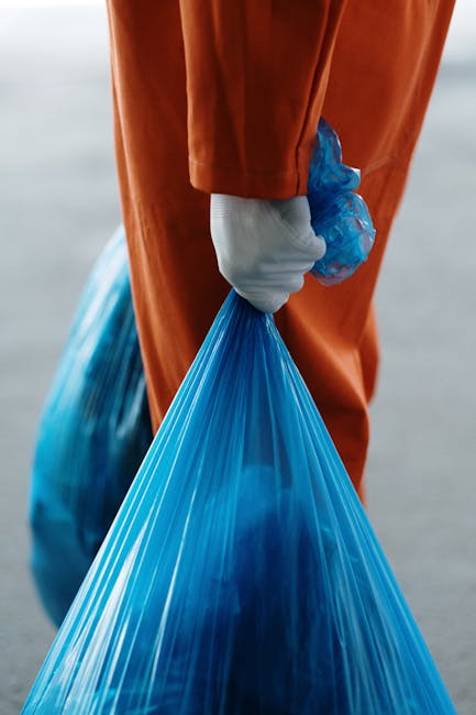 A close-up of a person wearing orange protective overalls and a white glove, holding a large, semi-transparent blue plastic bag filled with waste material. The individual is gripping the bag's gathere